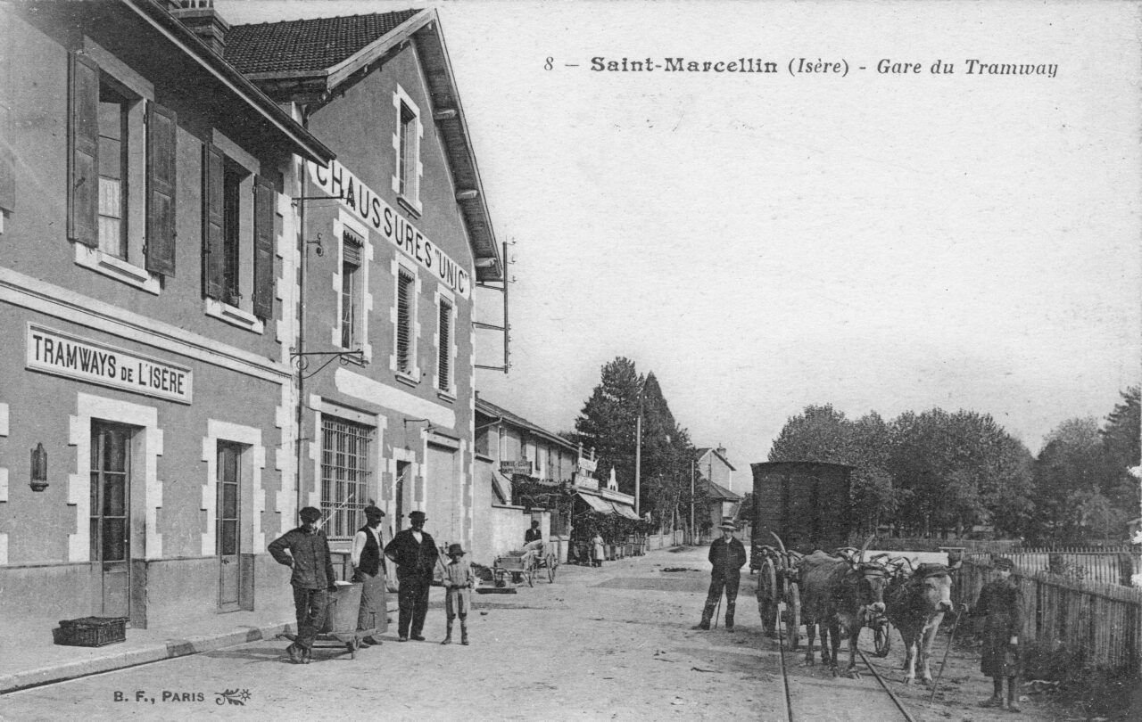 Carte postale ancienne en noir et blanc : la gare du tramway de Saint-Marcellin, avec la plaque « TRAMWAYS DE L'ISÈRE » sur la façade, un attelage de bœufs tirant une charrette le long des rails et un groupe d'hommes posant sur le quai.