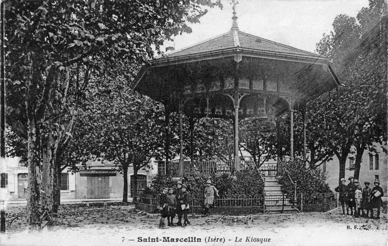 Carte postale ancienne en noir et blanc : le kiosque à musique de Saint-Marcellin, structure octogonale à colonnes de fonte et toit en pavillon, entouré de platanes et d'une grille basse, avec un groupe d'enfants posant devant.