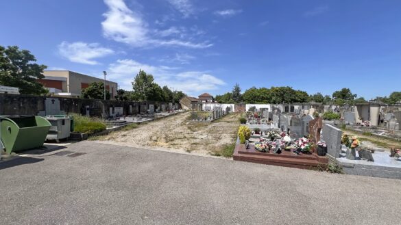 Entrée du cimetière de Saint-Marcellin, montrant une intersection d'allées bitumées plus propres au premier plan, s'ouvrant sur une large allée secondaire en friche en arrière-plan. Des monuments funéraires et un mur d'enceinte sont visibles sous un ciel bleu clair.