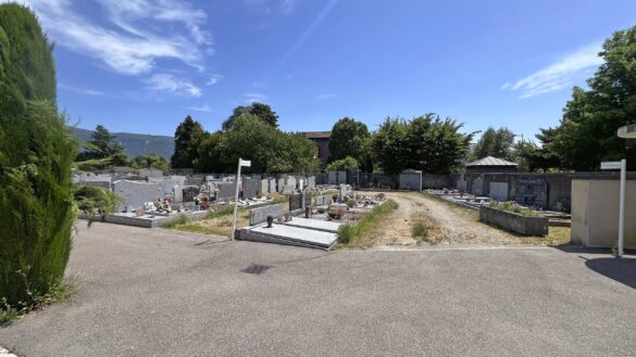 Plan moyen de tombes et monuments funéraires au cimetière de Saint-Marcellin, montrant de l'herbe sèche et des plantes sauvages poussant autour des sépultures et d'un poteau de signalisation.