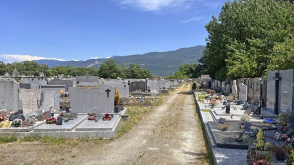 Vue en perspective d'une longue allée de gravier non entretenue et herbeuse, encadrée de chaque côté par des rangées de tombes en granit au cimetière de Saint-Marcellin, avec des montagnes boisées en arrière-plan sous un ciel bleu.